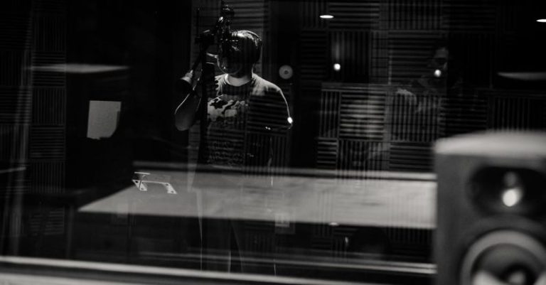 Voice Control - Through glass of black and white anonymous male musician in headphones singing with microphone while recording song in modern studio