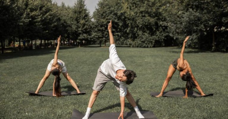 Flexible Displays - Group of diverse friends doing Revolved Wide Legged Forward Bend while exercising yoga on lawn